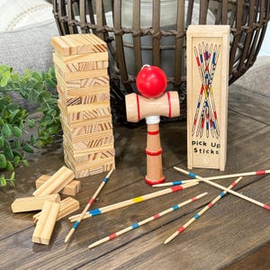 Op de afbeelding: Houten tafelspellen, waaronder een toren van blokken, een kendama-speelgoed met een rode bal en een doosje met plukstokken. De plukstokken zijn beschilderd met rode, blauwe en gele strepen. De doos heeft de tekst "pick Up Sticks".
