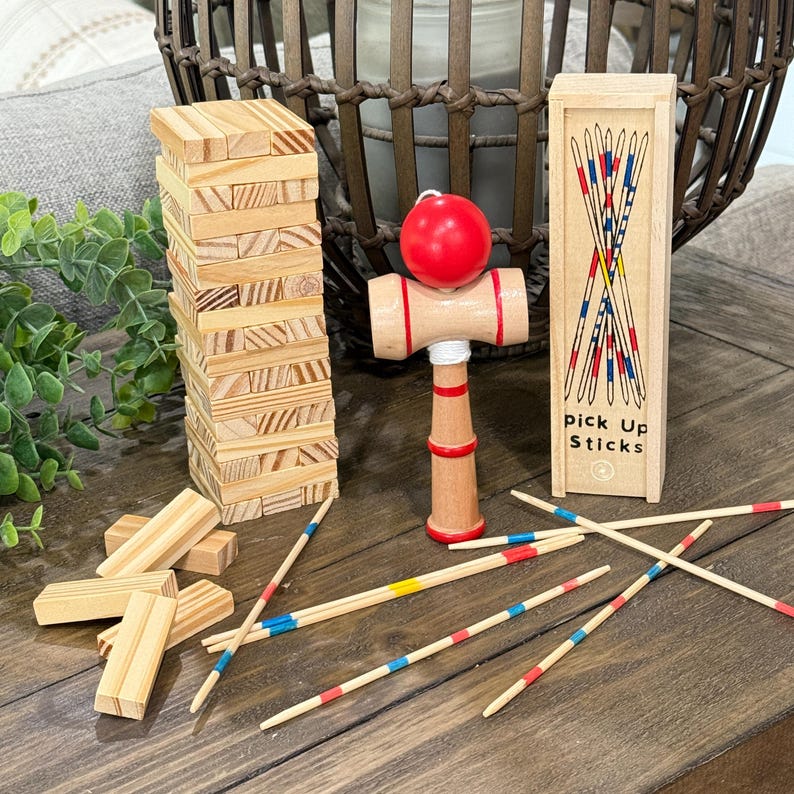 Op de afbeelding: Houten tafelspellen, waaronder een toren van blokken, een kendama-speelgoed met een rode bal en een doosje met plukstokken. De plukstokken zijn beschilderd met rode, blauwe en gele strepen. De doos heeft de tekst "pick Up Sticks".