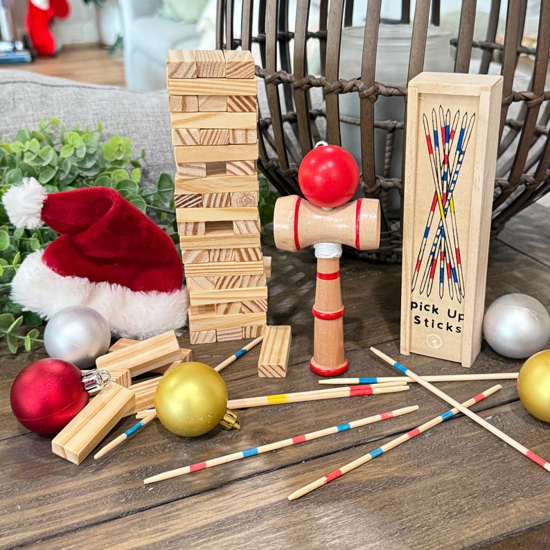 Op de afbeelding: Een verzameling houten speelgoed en kerstversieringen op een houten oppervlak. Inclusief een Jenga-toren, een kendama, een doosje Mikado en rode, gouden en zilveren ballen. Er is ook een rode kerstmuts aanwezig.