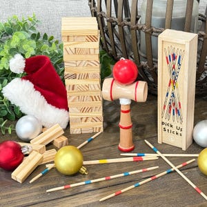 Op de afbeelding: Een feestelijke opstelling met een rode kerstmuts, een houten blokkentoren en een houten kendama-speelgoed met een rode bal. Bevat ook een doosje Mikado en kerstballen. De items zijn gerangschikt op een houten oppervlak.