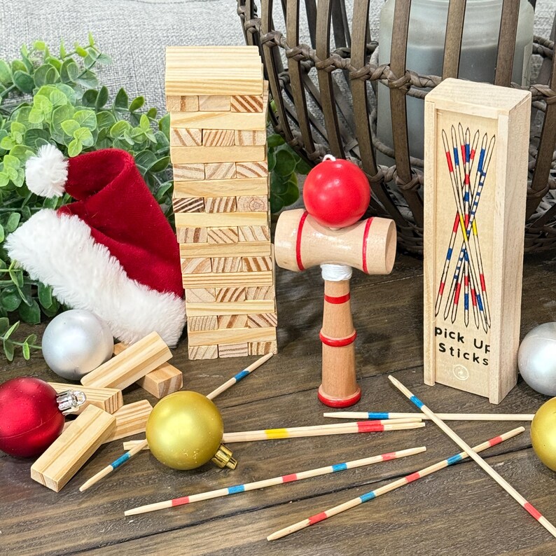 Op de afbeelding: Een feestelijke opstelling met een rode kerstmuts, een houten blokkentoren en een houten kendama-speelgoed met een rode bal. Bevat ook een doosje Mikado en kerstballen. De items zijn gerangschikt op een houten oppervlak.