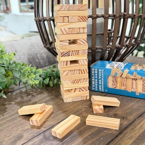 Op de afbeelding: Een houten tumbling tower game met een toren van rechthoekige blokken. Verschillende blokken liggen verspreid rond de toren. De speldoos is blauw met witte tekst "Tumbling Tower Game".