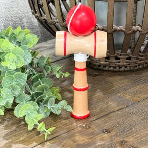Op de afbeelding: Een houten kendama-speelgoed met een rode bal die met een wit touwtje is bevestigd. De houten handgreep heeft rode strepen en accenten. Het speelgoed staat op een houten oppervlak met groen gebladerte op de achtergrond.