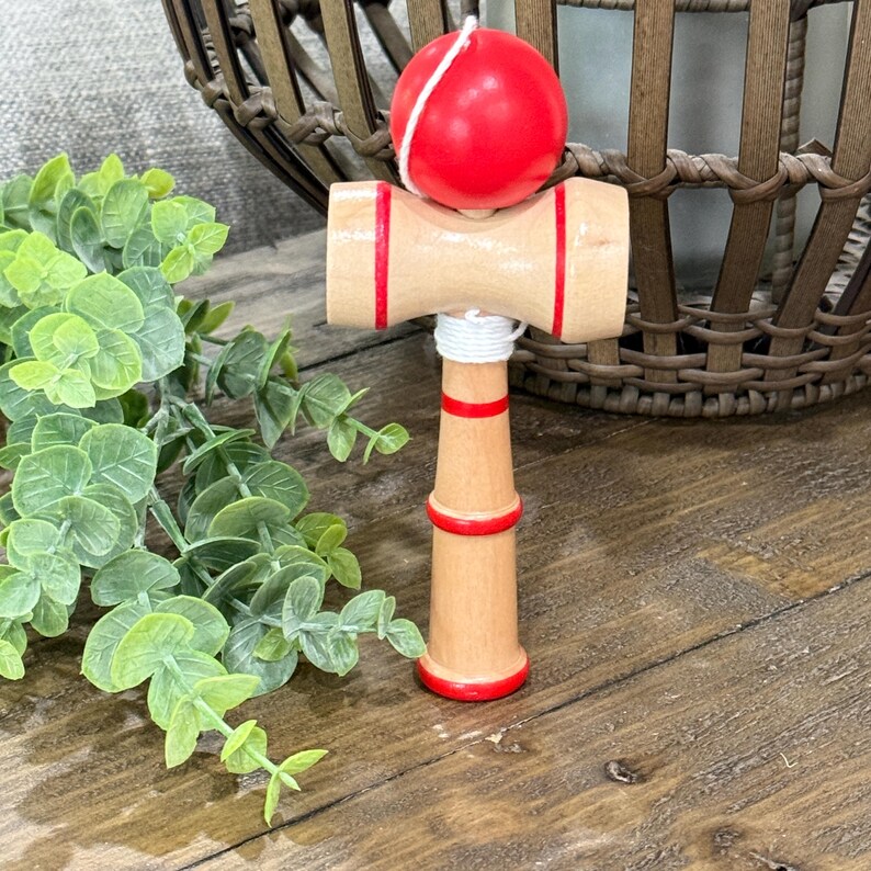 Op de afbeelding: Een houten kendama-speelgoed met een rode bal die met een wit touwtje is bevestigd. De houten handgreep heeft rode strepen en accenten. Het speelgoed staat op een houten oppervlak met groen gebladerte op de achtergrond.