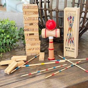 Op de afbeelding: Een selectie van houten spellen, waaronder een toren van houten blokken, een kendama-speelgoed met een rode bal en een doosje mikadostokjes. De mikadostokjes zijn rood, blauw en geel. Op de doos staat "pick up sticks".