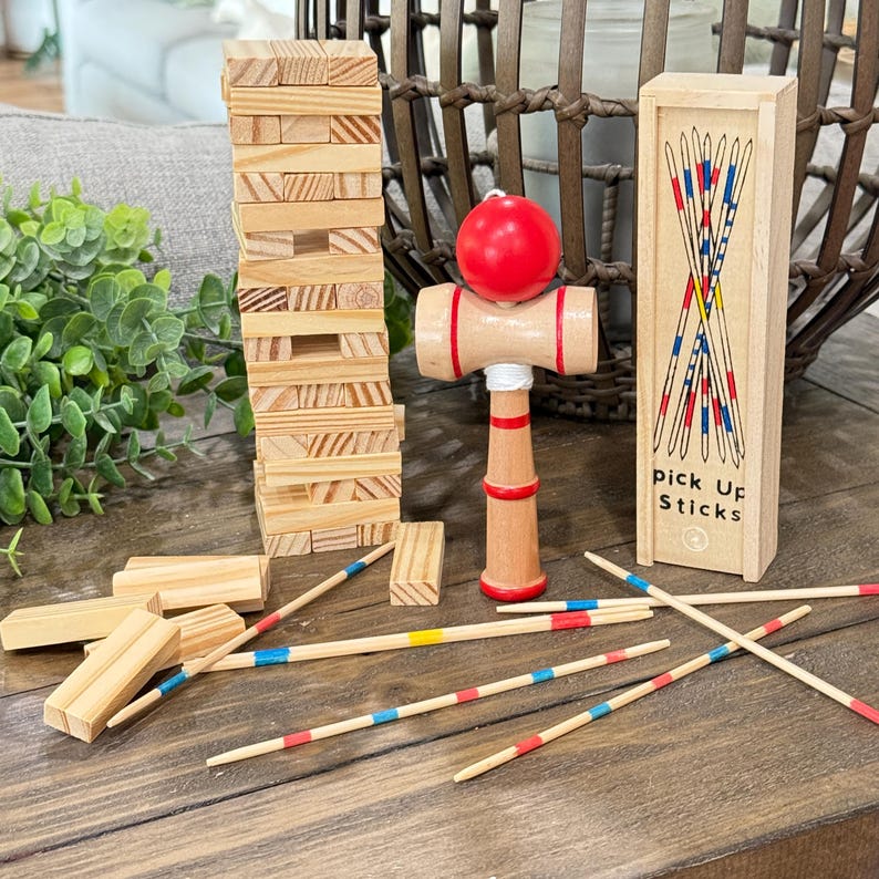 Op de afbeelding: Een selectie van houten spellen, waaronder een toren van houten blokken, een kendama-speelgoed met een rode bal en een doosje mikadostokjes. De mikadostokjes zijn rood, blauw en geel. Op de doos staat "pick up sticks".
