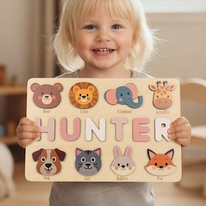 May include: A wooden name puzzle with the name "HUNTER" in pink letters. The puzzle features animal shapes including a bear, lion, elephant, giraffe, dog, cat, rabbit, and fox. The child is holding the puzzle.