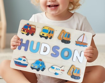 May include: Wooden puzzle featuring the name "HUDSON" in blue and white letters, surrounded by colorful vehicle shapes. Includes a fire truck, school bus, excavator, sailboat, ship, car, airplane, and another excavator.