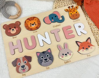 Puede incluir: Rompecabezas de madera con el nombre "HUNTER" en letras rosas y blancas, rodeado de piezas con forma de animales. Los animales incluyen un oso, un león, un elefante, una jirafa, un zorro, un perro, un gato y un conejo. El rompecabezas está sobre una tabla de madera clara.