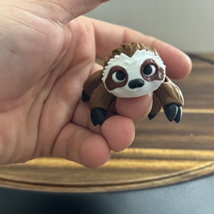 May include: A small, brown and white sloth figurine with black eyes and claws. The figurine is made of a soft, pliable material and is about the size of a person's hand.