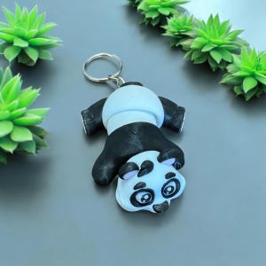 Könnte beinhalten: Ein Schlüsselanhänger in Form eines schwarz-weißen Pandas mit einem silbernen Schlüsselring. Der Panda liegt auf dem Rücken, die Pfoten in der Luft und der Kopf zur Seite geneigt.