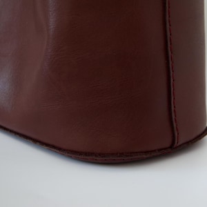 May include: Close-up of a burgundy leather tote bag. The bag's corner is visible, showcasing the smooth texture of the leather and the red stitching along the seams. The bag sits on a white surface.