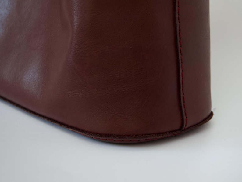 May include: Close-up of a burgundy leather tote bag. The bag's corner is visible, showcasing the smooth texture of the leather and the red stitching along the seams. The bag sits on a white surface.
