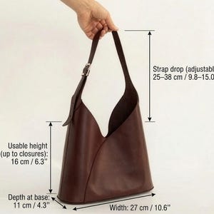 May include: A dark brown leather tote bag with a unique, open design. The bag has a strap drop of 9.8-15.0 inches, a usable height of 6.3 inches, a depth at the base of 4.3 inches, and a width of 10.6 inches. The strap is adjustable.