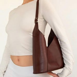 May include: A brown leather shoulder bag with a unique, open design. The bag has a long, adjustable strap with a silver buckle. The bag is held by a person wearing a white long-sleeve shirt and white pants.