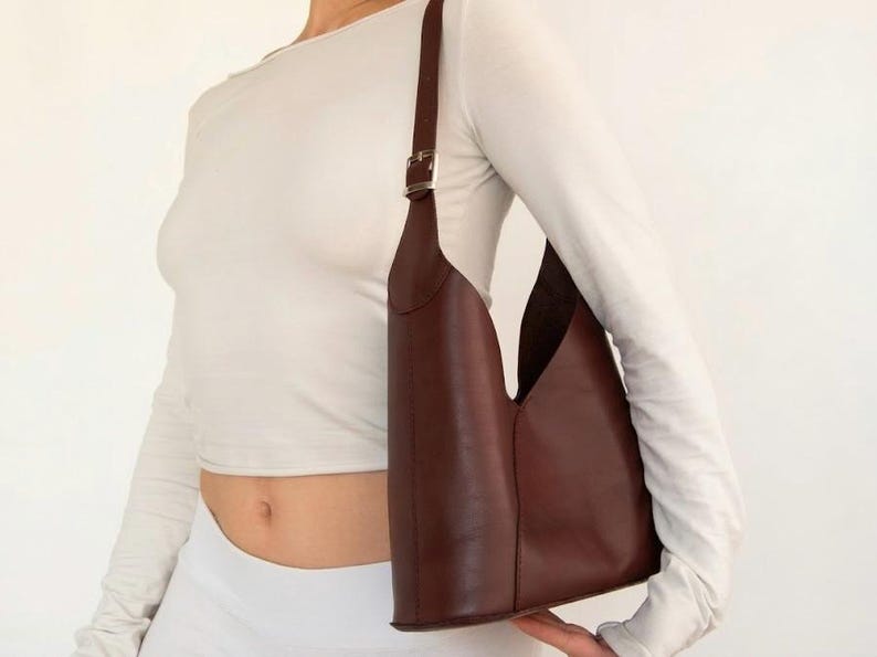 May include: A brown leather shoulder bag with a unique, open design. The bag has a long, adjustable strap with a silver buckle. The bag is held by a person wearing a white long-sleeve shirt and white pants.