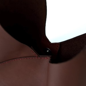 May include: Close-up of a burgundy leather bag with a magnetic closure. The bag's interior is visible, revealing a darker shade of burgundy. Red stitching accents the seams, adding a contrasting detail to the design.