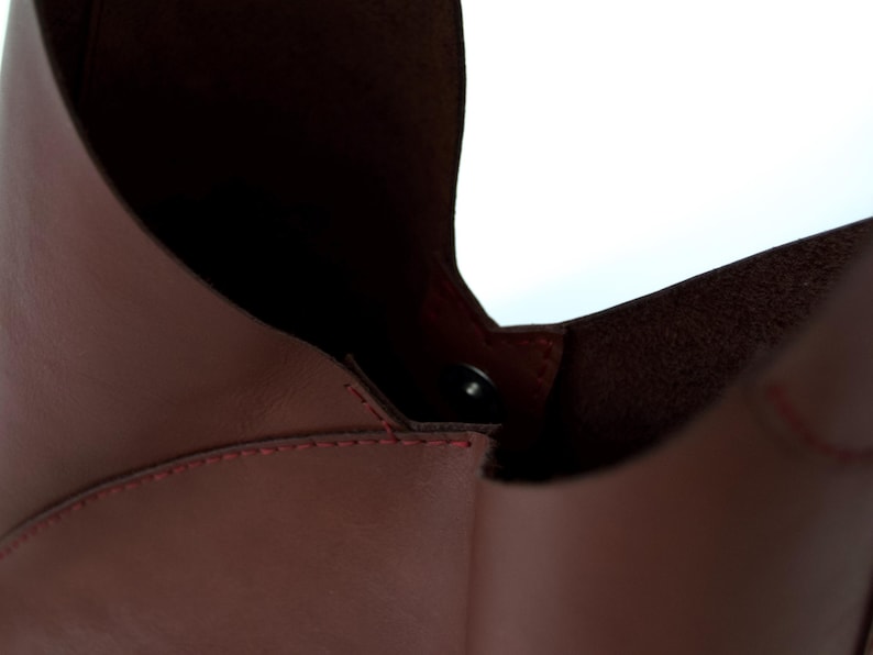 May include: Close-up of a burgundy leather bag with a magnetic closure. The bag's interior is visible, revealing a darker shade of burgundy. Red stitching accents the seams, adding a contrasting detail to the design.