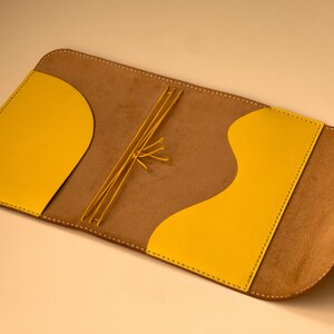 Puede incluir: Una cubierta de cuaderno de cuero marrón con detalles en cuero amarillo. La cubierta tiene una solapa que se cierra con una correa de cuero amarilla.