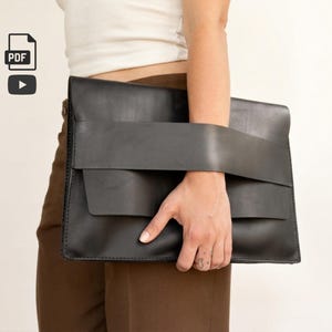 Peut inclure: Une pochette en cuir noir au design unique. Elle est dotée d'une large sangle pour le poignet et d'une fermeture à rabat. Le sac est tenu par une personne portant un haut blanc et un pantalon marron.