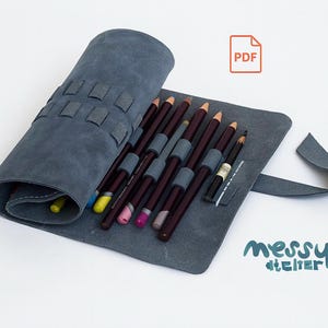 Puede incluir: Un estuche enrollado de cuero azul pizarra con cierre de solapa, que contiene varios lápices de colores. Los lápices son de varios colores y el estuche tiene un aspecto texturizado. El logotipo "messy atelier" es visible.