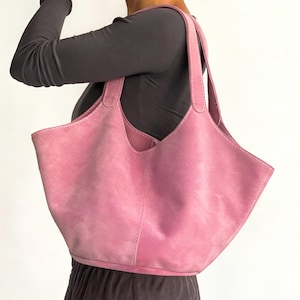 Puede incluir: Un bolso tote rosa con una parte superior ancha y abierta y dos asas. El bolso está hecho de un material suave y texturizado y presenta costuras blancas a lo largo de las costuras. El bolso se lleva al hombro.