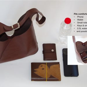 May include: A brown leather shoulder bag with a silver buckle, a wallet, a phone, and a wallet with a bird design. A water bottle is also present. The bag fits a phone, wallet, small notebook, keys, and a 0.5L water bottle.