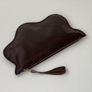 May include: A brown leather cloud-shaped clutch with a gold zipper and a brown leather strap.