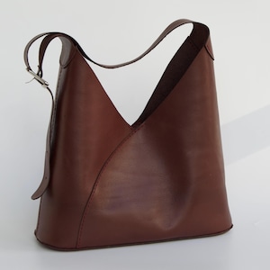 May include: A brown leather shoulder bag with a unique, angular design. The bag features a single shoulder strap with a silver buckle and a distinctive V-shaped opening. The bag is made of smooth, high-quality leather.