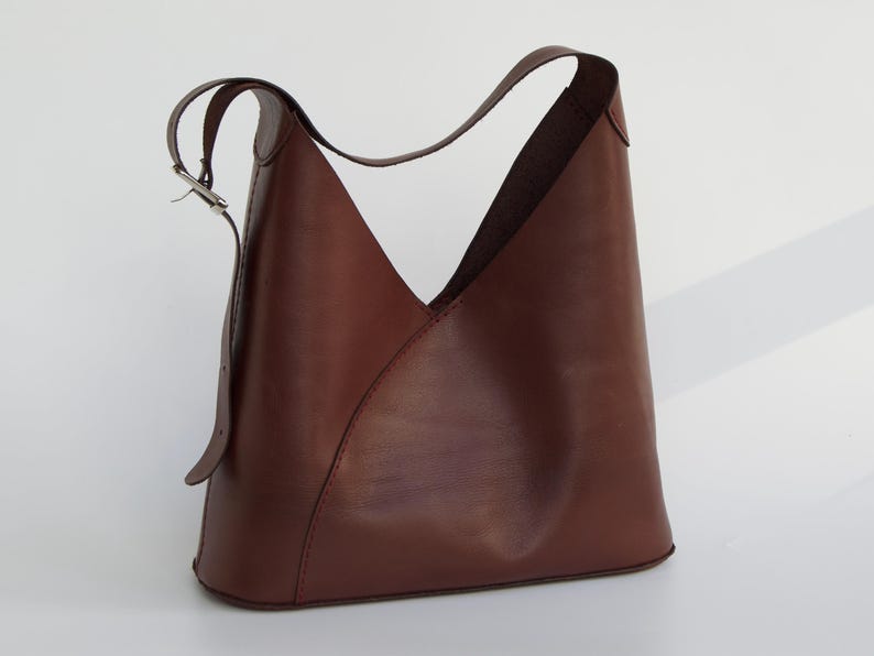 May include: A brown leather shoulder bag with a unique, angular design. The bag features a single shoulder strap with a silver buckle and a distinctive V-shaped opening. The bag is made of smooth, high-quality leather.
