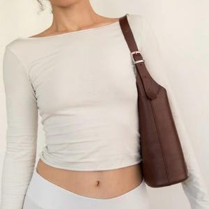 May include: A brown leather shoulder bag with a silver buckle on the strap. The bag is a dark brown color and has a sleek, minimalist design. The strap is adjustable. The bag is worn over the shoulder.