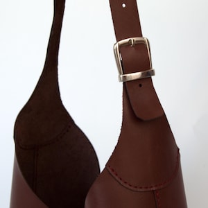 May include: Close-up of a brown leather bag with a silver buckle. The bag has a unique shape and design, with visible stitching and a strap. The leather appears to be high-quality.