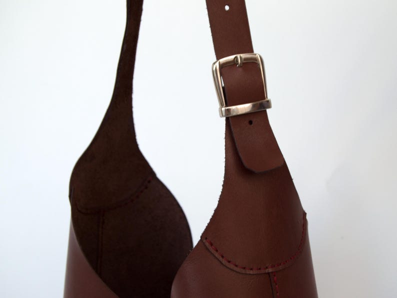 May include: Close-up of a brown leather bag with a silver buckle. The bag has a unique shape and design, with visible stitching and a strap. The leather appears to be high-quality.