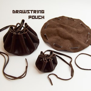 Può includere: Tre sacchetti con coulisse in pelle marrone scuro di varie dimensioni. Due sacchetti sono chiusi con cordini in pelle, mentre uno è steso in forma circolare con occhielli metallici e un cordino infilato. Il testo DRAWSTRING POUCH è visibile.