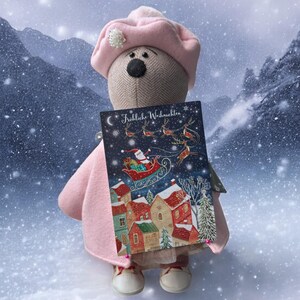 May include: A soft toy bear in a pink coat and hat, holding a Christmas card. The card depicts a winter scene with Santa's sleigh, reindeer, and snow-covered houses. The card reads "Fröhliche Weihnachten".