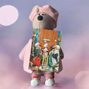 Könnte beinhalten: Ein rosa Stofftierbär mit einem rosa Barett hält eine Karte mit einer Illustration von zwei Frauen, die vor einer Tür mit Briefkästen stehen. Die Karte zeigt eine braune Holztür mit einem Messing-Türgriff und einem Briefkasten, aus dem ein weißer Briefumschlag herausragt.