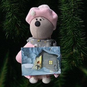 Könnte beinhalten: Handgefertigte Stoffpuppe mit rosa Barett, die eine kleine gerahmte Illustration einer Winterszene mit einer Hütte und einem Zwerg hält. Die Puppe hat einen grau gemusterten Körper, eine schwarze Nase und einen grauen Schal mit floralen Akzenten.