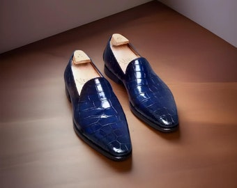 May include: A pair of navy blue leather loafers with a crocodile skin texture. The shoes have a low heel and a rounded toe. Wooden shoe trees are inserted inside the shoes. The shoes are displayed on a brown surface.