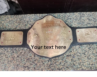 May include: A gold-colored wrestling championship belt with the words "WORLD HEAVYWEIGHT WRESTLING CHAMPION" engraved on the center plate. The belt has a black leather strap and red jewel accents.