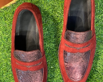 Könnte beinhalten: Ein Paar bordeauxfarbene Wildleder-Loafer mit einem metallischen, strukturierten Blatt. Die Schuhe haben ein klassisches Slip-on-Design mit dunklem Innenraum. Die Loafer werden auf einem Bett aus grünem Kunstgras präsentiert.