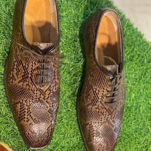 Puede incluir: Un par de zapatos de vestir de cuero marrón con un estampado de piel de serpiente. Los zapatos tienen cordones y están sobre una superficie verde. Los zapatos tienen un estilo Oxford clásico.