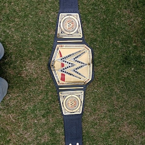 Puede incluir: Un cinturón de campeonato de lucha libre negro y dorado con el logotipo de la WWE. El cinturón tiene una placa central grande y dos placas laterales más pequeñas, todas con detalles dorados. El cinturón es de cuero negro y está sobre hierba verde.