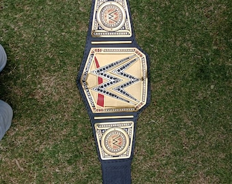 May include: A black and gold wrestling championship belt with the WWE logo. The belt has a large central plate and two smaller side plates, all with gold accents. The belt is made of black leather and is lying on green grass.