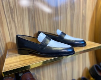 May include: A pair of black and white leather loafers displayed on a wooden shelf. The shoes have a classic design with a black body and a white vamp. The shelf has a gold trim.