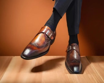 May include: Brown leather dress shoes with a single monk strap buckle closure. The shoes have brogue detailing and a polished finish. The image shows the shoes from a low angle, highlighting their design and craftsmanship. The shoes are part of a formal footwear collection.