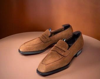 Könnte beinhalten: Ein Paar braune Penny-Loafer aus Wildleder mit schwarzen Sohlen, präsentiert auf einer hellbraunen Oberfläche. Die Schuhe haben ein klassisches Design mit einem Penny-Riemen-Detail. Die Loafer sind aus Wildleder.