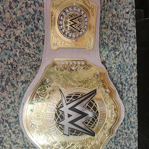 May include: A gold and silver championship belt with the WWE logo. The belt has a light pink leather strap and is adorned with intricate gold detailing and sparkling embellishments. The words "CHAMPION" are visible.