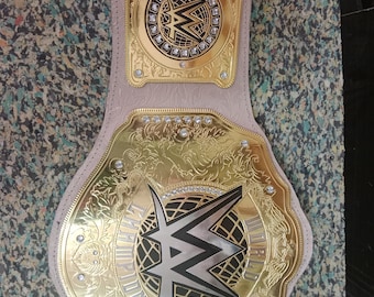 May include: A gold and silver championship belt with the WWE logo. The belt has a light pink leather strap and is adorned with intricate gold detailing and sparkling embellishments. The words "CHAMPION" are visible.