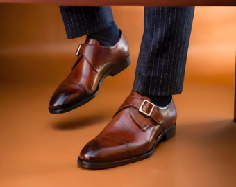 May include: A pair of brown leather monk strap shoes with gold buckles. The shoes have a polished finish and are set against a warm, orange-brown background. The image shows the shoes from a low angle.