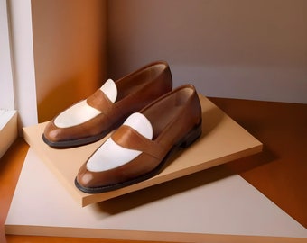 May include: A pair of brown leather loafers with white vamps. The shoes are displayed on a beige platform against a warm-toned background. The classic design features a slip-on style.